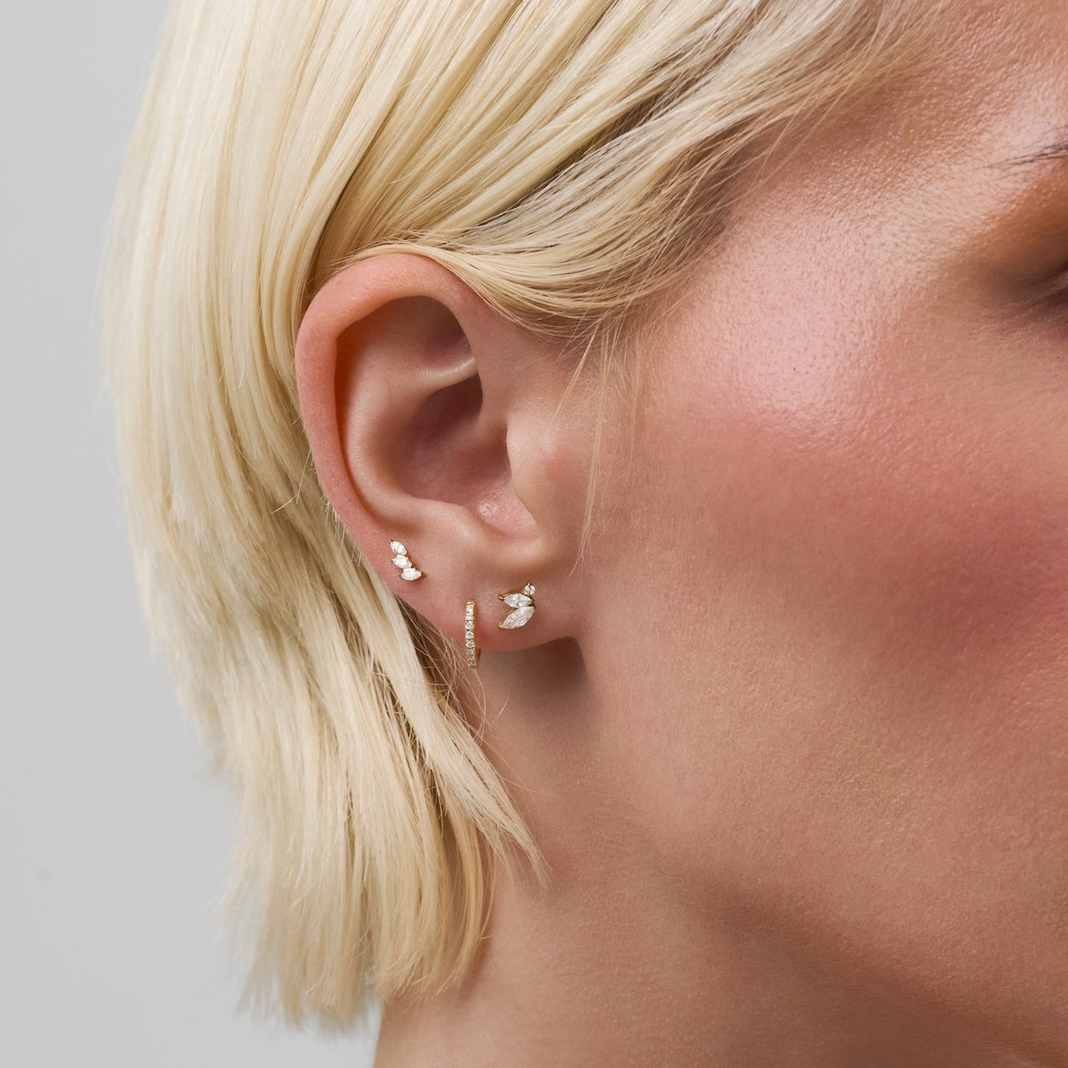 Close-up of a model's ear wearing multiple delicate gold earrings, including small curved bar studs and a small vertical huggie hoop, against a light gray background.